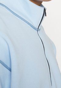 Světle modrý pulovr na zip s vysokým límcem, se hladkou texturou a jemným kontrastním prošíváním podél švů.