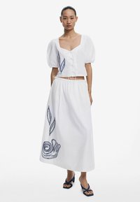 Witte katoenen tweedelige outfit met een cropped top met ballonmouwen en een lange rok versierd met blauwe bladeren en spiraal borduursels.