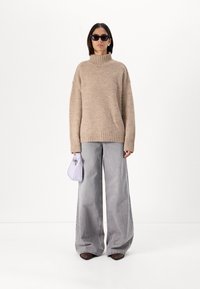Beige knitted sweater with high neck, paired with light gray wide-leg jeans. Model holds a small lavender handbag and wears black sunglasses.