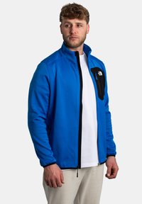 Veste softshell bleue avec une poche poitrine zipp ée noire, fermeture éclair sur le devant et poignets élastiques. Le matériau semble lisse et légèrement texturé.