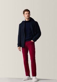 Marineblauer Peacoat über einem marineblauen Kabelstrickpullover, kombiniert mit roten Slim-Fit-Hosen und weißen Sneakers. Glatte Stoffe, maßgeschneiderte Passform und kontrastierende Texturen.