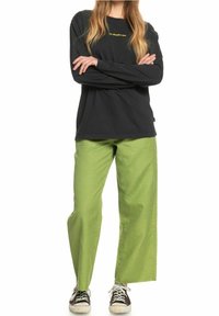 Svart långärmad skjorta med gult logotyp, kombinerad med löst sittande gröna byxor. Modellen bär svarta och vita sneakers.