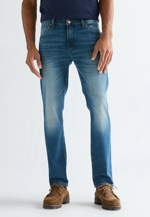 Mann trägt blaue ausgebleichte Jeans und braune Schnürschuhe, steht vor einem schlichten hellen Hintergrund.