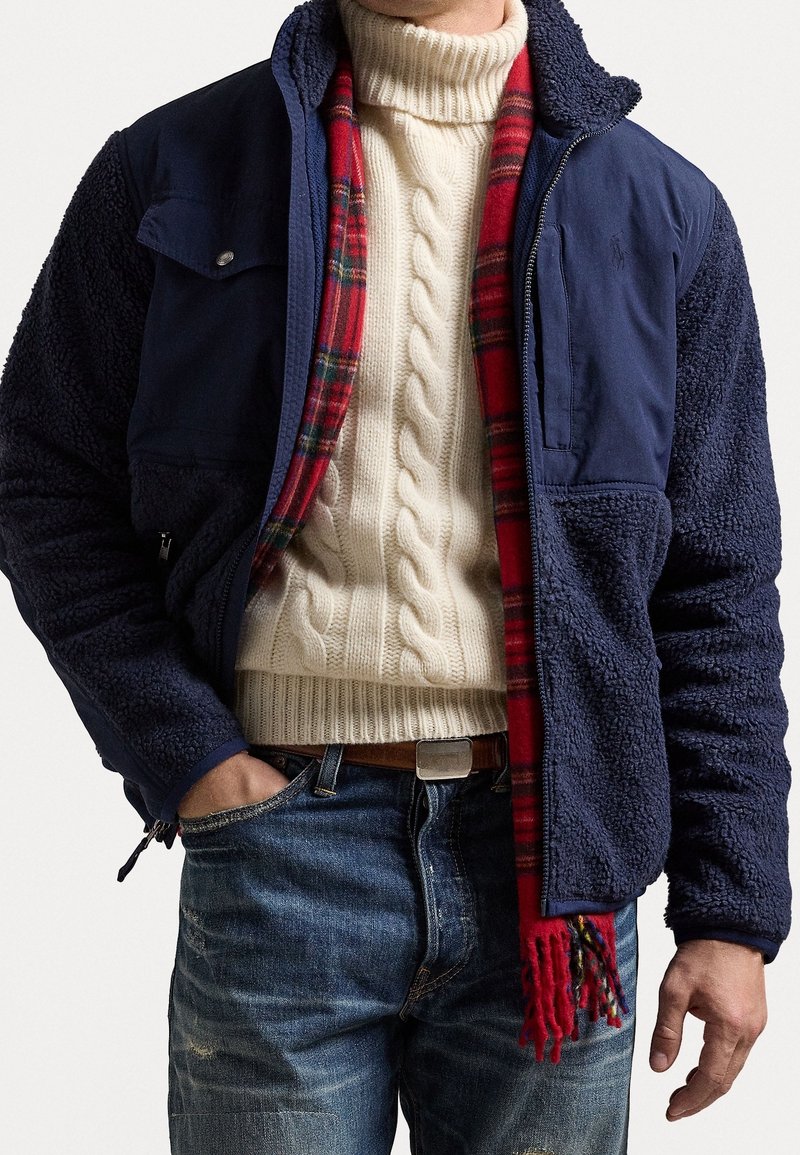 Marineblaue Fleecejacke mit strukturierter Oberfläche, mit Reißverschluss versehenen Taschen und einem karierten rot-schwarzen Schal; getragen über einem cremefarbenen Kabelstrickpullover.