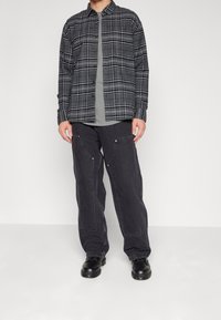 Camisa de cuadros negra y gris sobre una camiseta gris, combinada con jeans negros holgados con bolsillos angulados y zapatos negros.