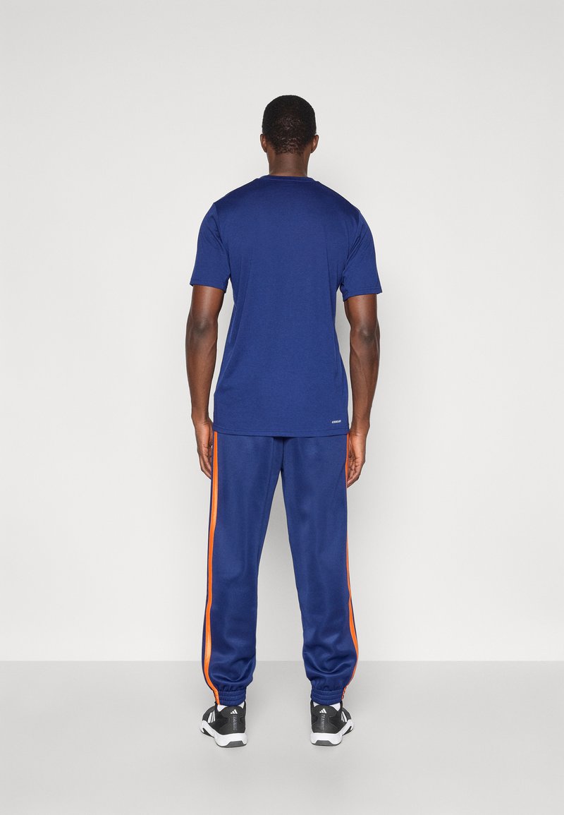 Navyblaues T-Shirt und Sweatpants mit orangefarbenen Seitenstreifen, aus weichem Stoff gefertigt. Verfügt über elastische Bündchen und einen lockeren Schnitt. Sportliche Schuhe sind sichtbar.