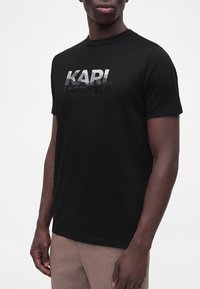 Άνδρας που φοράει μαύρο κοντομάνικο T-shirt με το "KARL LAGERFELD" τυπωμένο μπροστά σε μεταλλικό ασημί και μαύρο κείμενο με διαβάθμιση.