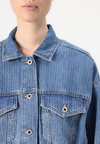 Giubbotto di denim blu chiaro con righe verticali, dotato di colletto a bollina, bottoni a pressione e due tasche sul petto con pattine.
