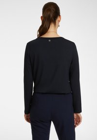 Námořnický modrý top s dlouhým rukávem a kulatým výstřihem, s jemným logem na zadní straně. Měkká tkanina s hladkým povrchem.