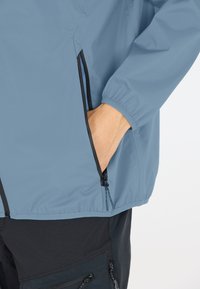 Lätt blå jacka med elastiska manschetter och sidofickor med svarta dragkedjor, tillverkad av slät, vattenavvisande tyg.