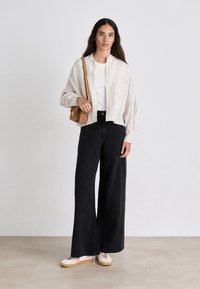 Camicia button-up beige a righe leggere, t-shirt bianca, pantaloni neri larghi, sneakers beige e una piccola borsa a tracolla marrone.