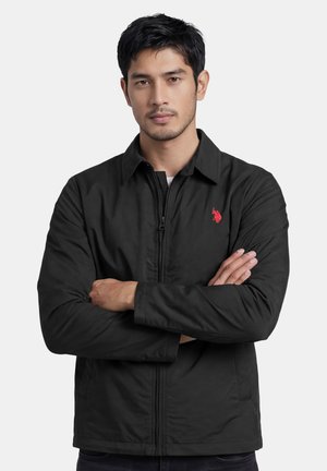 Mann mit verschränkten Armen, der eine schwarze Reißverschlussjacke mit kleinem rotem Logo auf der linken Brust vor einem einfarbigen hellen Hintergrund trägt.