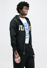 Čierna mikina na zips so zlatým nápisom, v kombinácii s bielym tričkom s modrým grafickým motívom a čiernymi teplákmi.