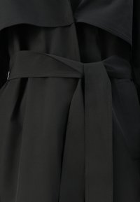 Trench coat in tessuto nero, aderente in vita con una cintura coordinata, che mostra dettagli di cucitura e un ampio bavero.