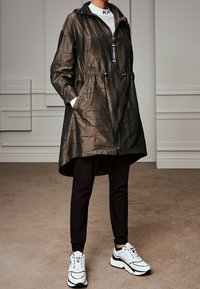 Parka impermeabile di colore verde oliva con cappuccio, vita cinchata e chiusura con zip. Presenta accenti neri a contrasto e dettagli del logo stampati.