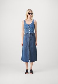 Abito in denim con parte superiore aderente e chiusura a bottoni, gonna ampia e spacco frontale. Colore: blu scuro. Abbinato a sandali neri scivolati.