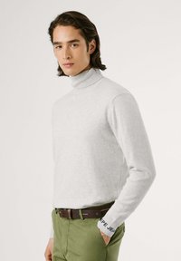 Maglione dolcevita grigio chiaro in tessuto morbido con design aderente, abbinato a pantaloni verdi e una cintura marrone. I polsini presentano un dettaglio con logo blu.