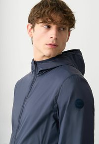 Marineblaue Kapuzenjacke mit glatter, glänzender Textur. Verfügt über einen durchgehenden Reißverschluss und ein rundes Logopatch auf der linken Schulter.