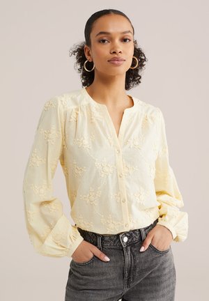 Frau mit lockigem Haar, die Creolen, eine blassgelbe, mit Blumen bestickte Bluse und schwarze, hochtaillierte Jeans trägt, mit den Händen in den Taschen.