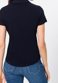 Žena v námořnicky modré košili s krátkými rukávy a modrých džínách, zobrazená zezadu na jednobarevném pozadí.