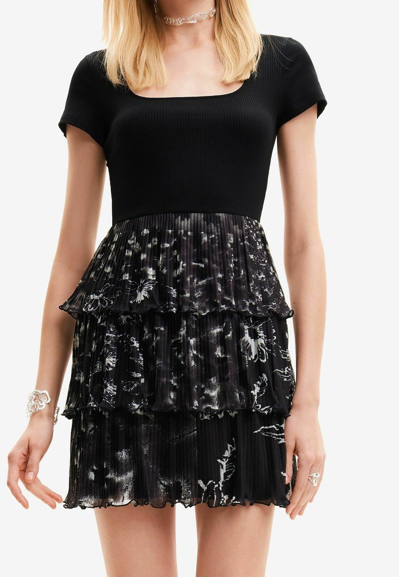 Femme portant une courte robe noire avec un haut côtelé et une jupe plissée à étages avec des motifs floraux blancs.