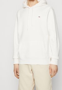 Persona vestida con un sudadera blanca con capucha y un pequeño logo de Tommy Hilfiger en el pecho, y pantalones beige, de frente.