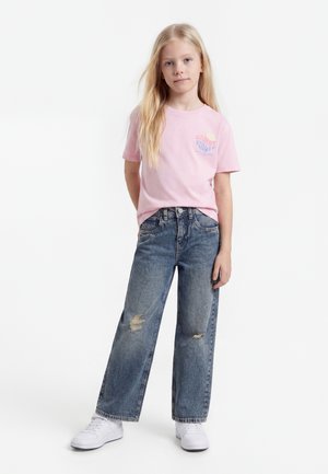Jeune fille aux longs cheveux blonds portant un t-shirt rose à motif, un jean bleu déchiré et des baskets blanches, debout devant un fond blanc.