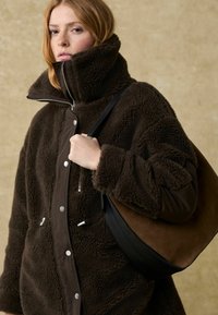 Bruine schapenvachtjas met hoge kraag, zilverkleurige hardware en zwarte accenten. Model houdt een bruin-en-zwarte schoudertas vast voor een neutrale achtergrond.