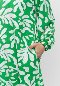 Vihreä mekko valkoisella abstraktilla lehtikuviolla, jossa on pitkät hihat joustavuuskuminauhoilla ja sivutasku. Sujuva, kevyt kangastuntuma.