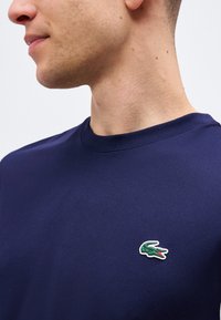 Man wearing a navy blue shirt with a small green crocodile logo on the left chest, shown from neck to mid-torso.