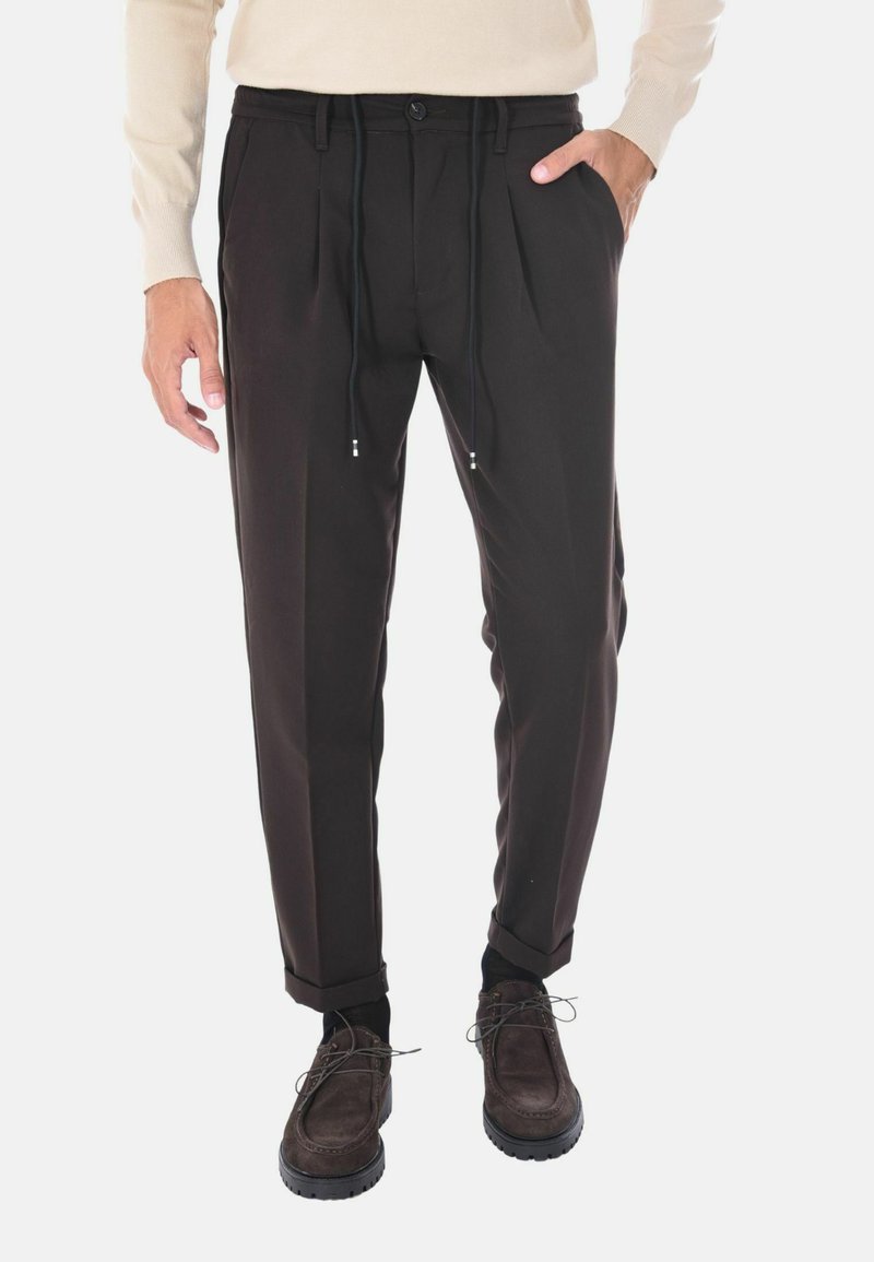 Pantaloni neri plissettati con vita elastica, chiusura con bottone e lacci; indossati con scarpe scure, creando un look casual ma raffinato.