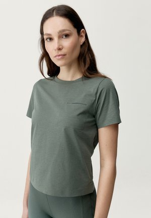 Kvinde med langt brunt hår iført en grøn kortærmet skjorte med brystlomme, kombineret med grønne ribstrikkede bukser mod en ensfarvet baggrund.