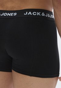 Melnas bokserbikses no mīksta auduma ar platu jostu, uz kuras rakstīts "JACK & JONES" baltā krāsā. Gluda tekstūra un piegulošs izmērs.