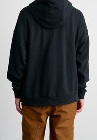 Černá mikina s kapucí s volnějším střihem, velkou kapucí, dlouhými rukávy a žebrovanými manžetami. Kombinována se světle hnědými kalhotami.