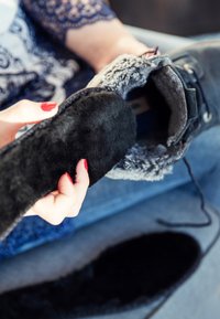 Black faux fur insole held above a dark boot with a gray fur cuff. Background shows blue jeans and a textured surface.
