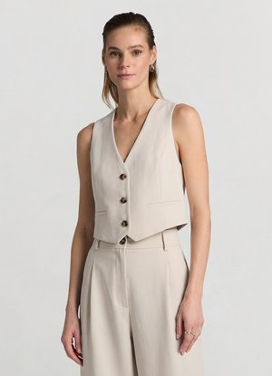 Donna che indossa un gilet beige senza maniche con tre bottoni e pantaloni a vita alta con gambe larghe abbinati su uno sfondo semplice.
