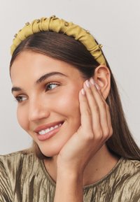 Yellow fabric headband with gathered texture and decorative gold accents, complemented by a model's neutral top and natural makeup.