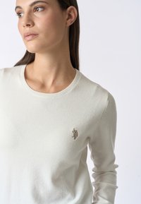 Maglione avorio chiaro con scollo rotondo e maniche lunghe. Presenta un emblema texturizzato sul lato sinistro del petto. Tessuto morbido con finitura liscia.