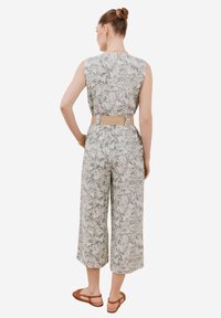 Ärmlös jumpsuit i ljusgrön med lövmönster, avskuren vida ben, åtdragen i midjan med ett beige bälte. Bruna sandaler syns.