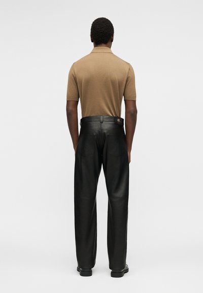 Άνδρας που φοράει κοντομάνικο μπεζ πόλο πουκάμισο, μπροστά σε απλό φόντο με γυρισμένη πλάτη, ενώ φορά ψηλόμεσα μαύρα δερμάτινα παντελόνια.