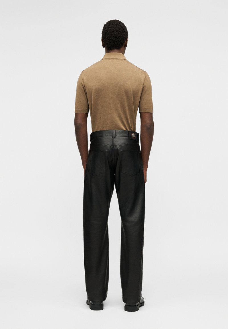 Homme portant un polo beige à manches courtes rentré dans un pantalon en cuir noir taille haute, debout de dos devant un fond uni.