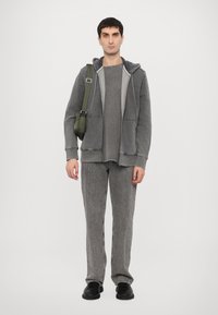 Grauer Hoodie mit einer Fronttasche, über einem passenden grauen Shirt getragen. Strukturierten, locker sitzenden grauen Hosen. Schwarze Schuhe und eine grüne Umhängetasche.