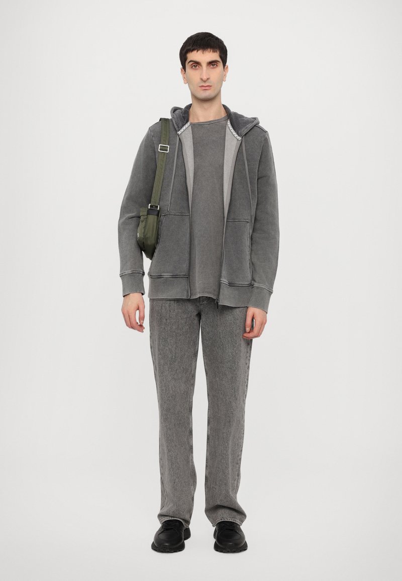 Grauer Hoodie mit einer Fronttasche, über einem passenden grauen Shirt getragen. Strukturierten, locker sitzenden grauen Hosen. Schwarze Schuhe und eine grüne Umhängetasche.