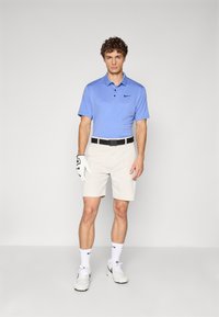Mandens blå polo shirt med knapper, beige shorts, sort bælte og hvid golfhandske. Hvide sokker og sneakers fuldender outfittet.