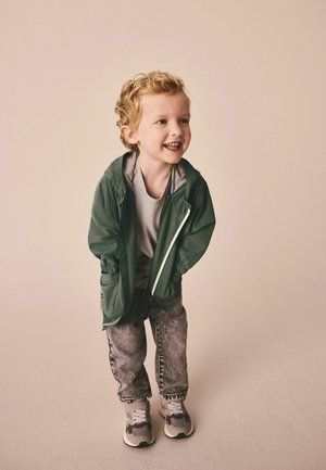 Grüne leichte Jacke mit grauen Akzenten, graues T-Shirt und ausgewaschene Jeans. Die Sneakers sind grau mit schwarzen Akzenten. Das Kind lächelt.
