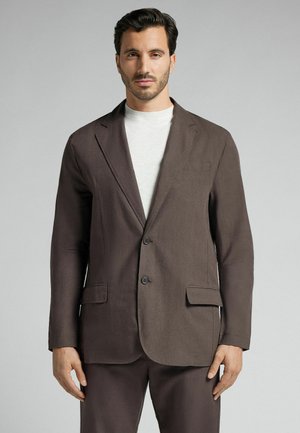 Brauner Leinenblazer mit Schalkragen, zwei Knöpfen und seitlichen Taschen. Kombiniert mit einem hellen, Rundhals-Pullover. Glatte Textur.