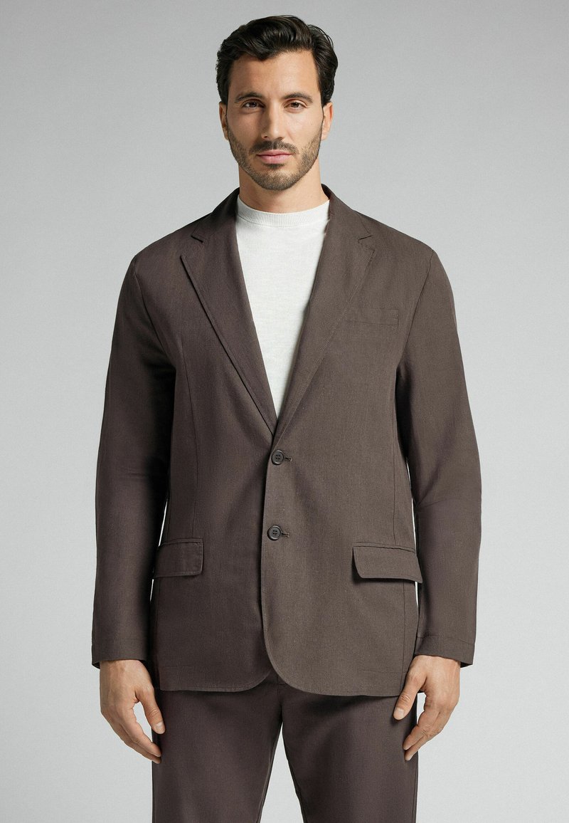 Brauner Leinenblazer mit Schalkragen, zwei Knöpfen und seitlichen Taschen. Kombiniert mit einem hellen, Rundhals-Pullover. Glatte Textur.