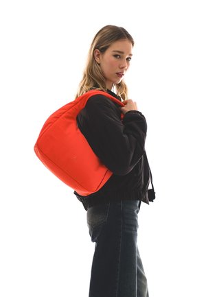 Tânără purtând o geacă neagră și blugi închiși la culoare, ținând o poșetă roșie aprinsă pe umăr, privind peste umăr pe fundal alb.