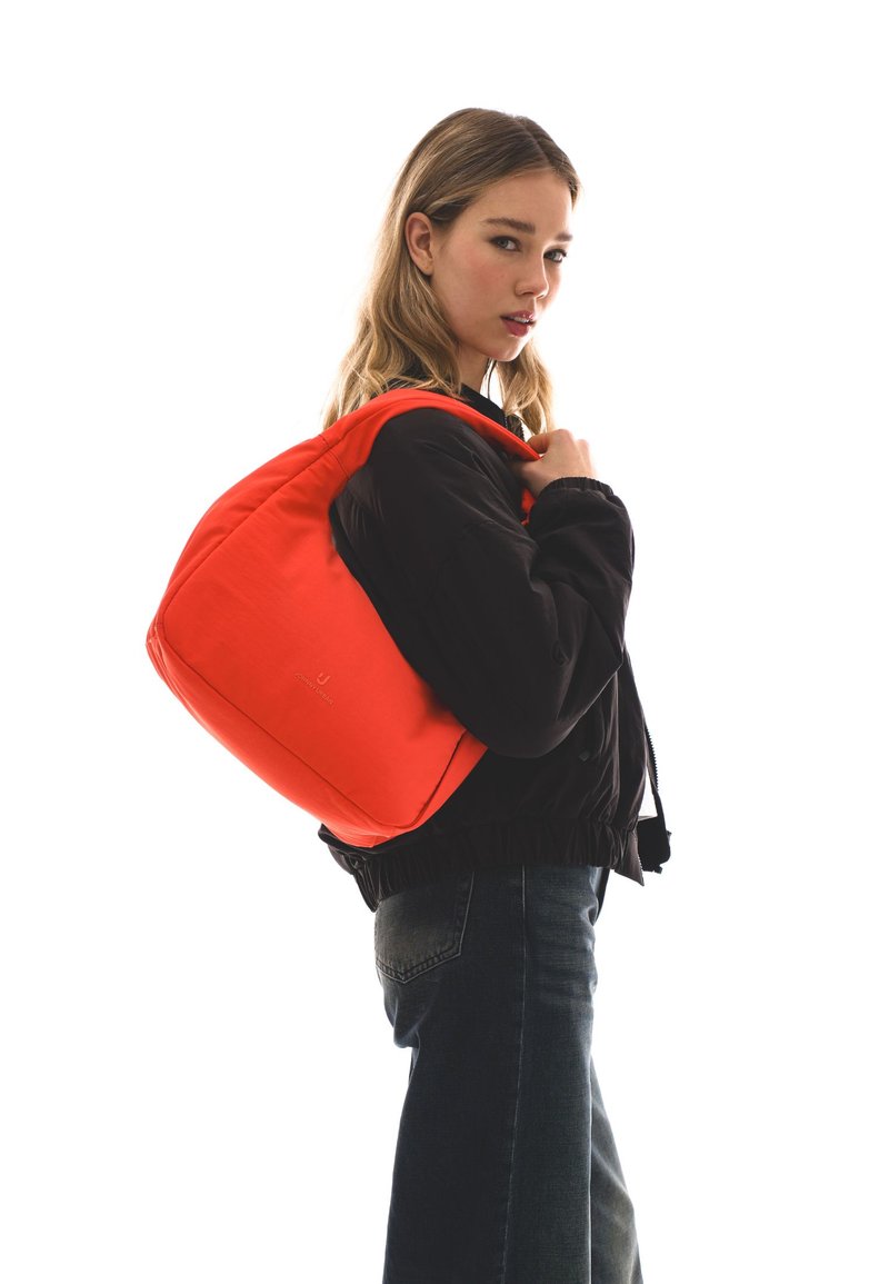 Fiatal nő fekete dzsekit és sötét farmert viselve, élénkpiros válltáskát tartva, hátrapillant fehér háttér előtt.