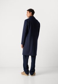 Abrigo de mezcla de lana de color navy con un diseño ajustado, silueta larga, cuello de muesca y puños abotonados, combinado con pantalones azul oscuro y zapatos beige.